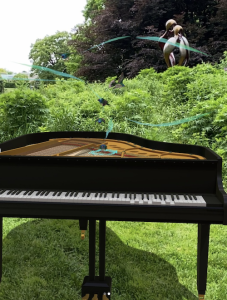 AR art, Dawn Chorus by Sarah Meyohas, as viewed through a smartphone. Viewer sees a grand piano in augmented reality with AR birds swooping around, bordered by a beautifully landscaped garden in real life.